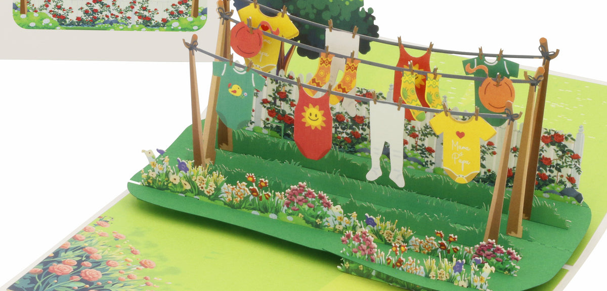 Aufgeklappte Pop-up-Karte mit Babywäsche und Stramplern auf einer Wäscheleine in einem blühenden Garten, dahinter die weiße Kartenvorderseite mit Wäscheleinen-Motiv.