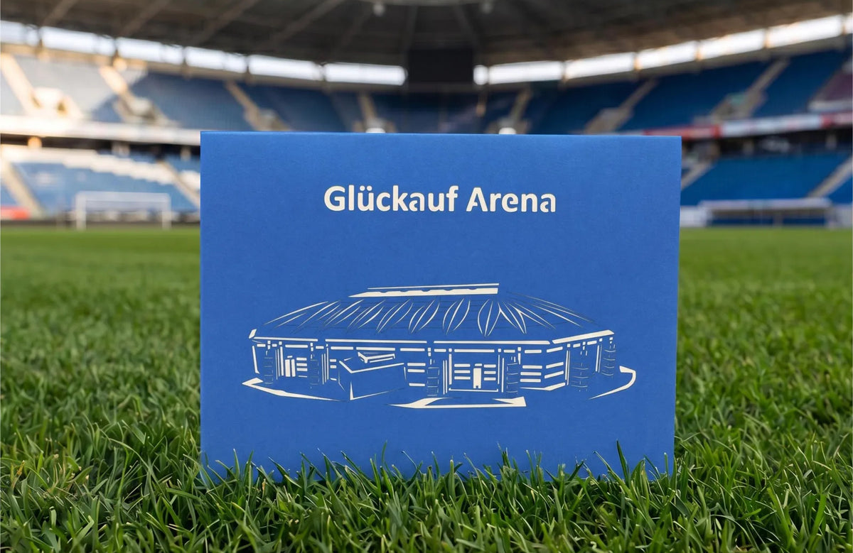 Blaue Klappkarte mit Scherenschnitt eines Fußballstadions und dem Schriftzug Glück auf Arena auf Rasen im Stadion.