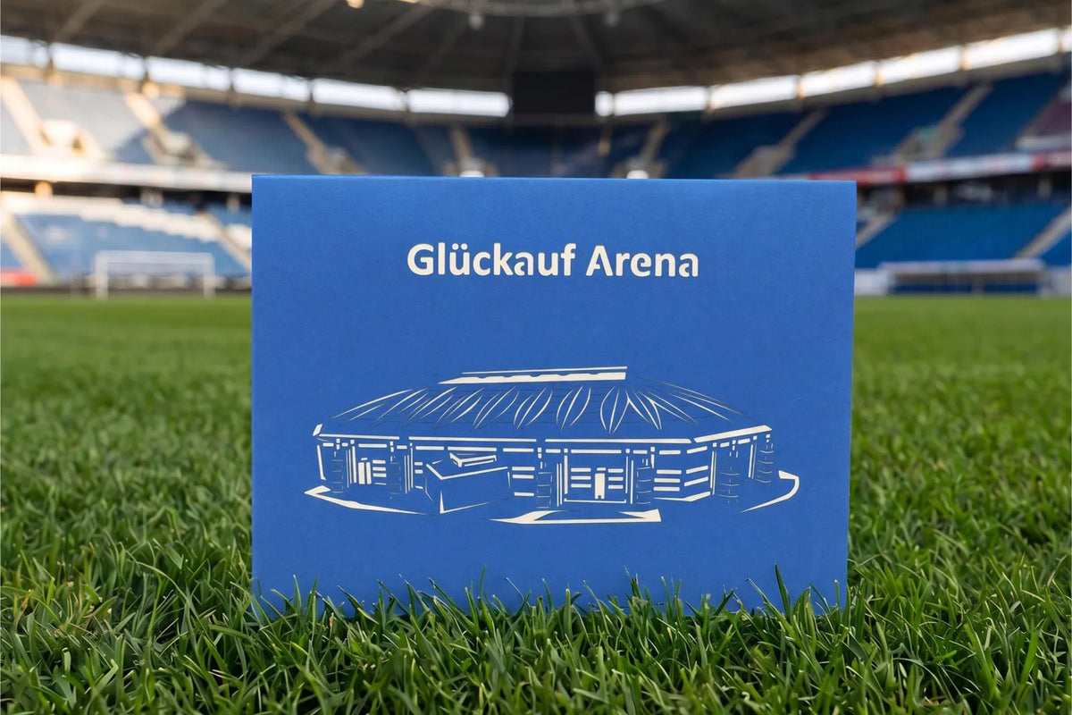 Blaue Klappkarte mit Scherenschnitt eines Fußballstadions und dem Schriftzug Glück auf Arena auf Rasen im Stadion.