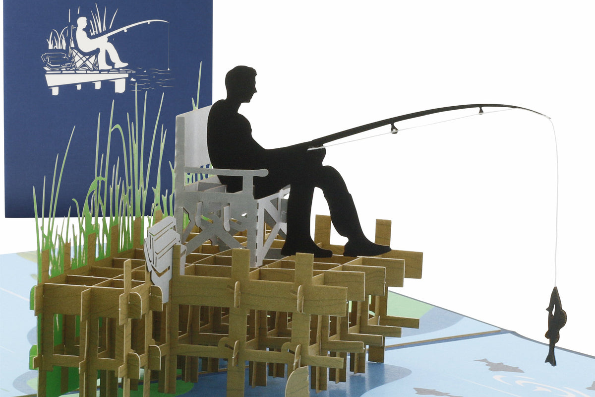 Pop-up-Karte mit einem Angler auf dem Steg, der auf einem Stuhl sitzt und angelt, im Hintergrund eine blaue Klappkarte mit einem Anglermotiv als Scherenschnitt.