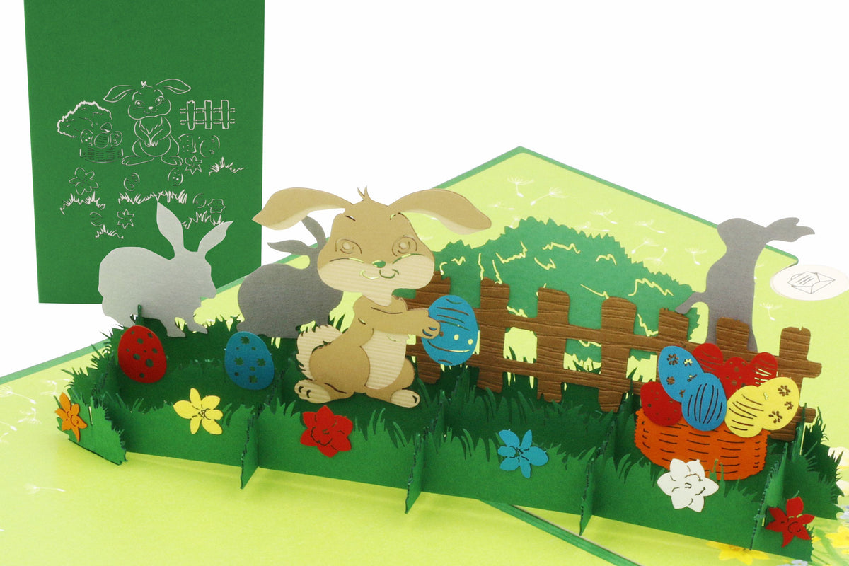 Aufgeklappte Pop-up-Karte mit Osterhasen, Ostereiern und Gartenzaun auf einer Frühlingswiese, daneben die grüne Kartenvorderseite mit Hasen-Scherenschnitt.