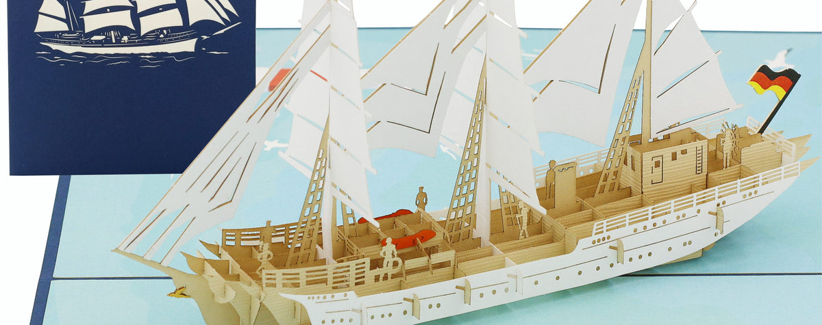 Aufgeklappte Pop-up-Karte mit dem Segelschiff Gorch Fock und Deutschlandflagge, dahinter die blaue Kartenvorderseite mit Segelschiff-Scherenschnitt.