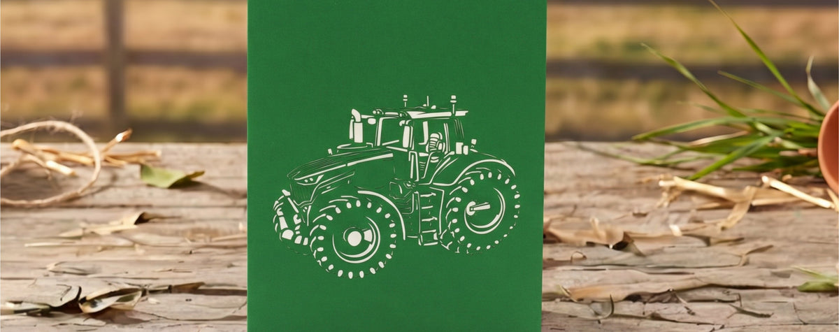 Grüne Klappkarte mit einem Scherenschnitt eines Traktors auf einem Holztisch vor unscharfem Bauernhofhintergrund