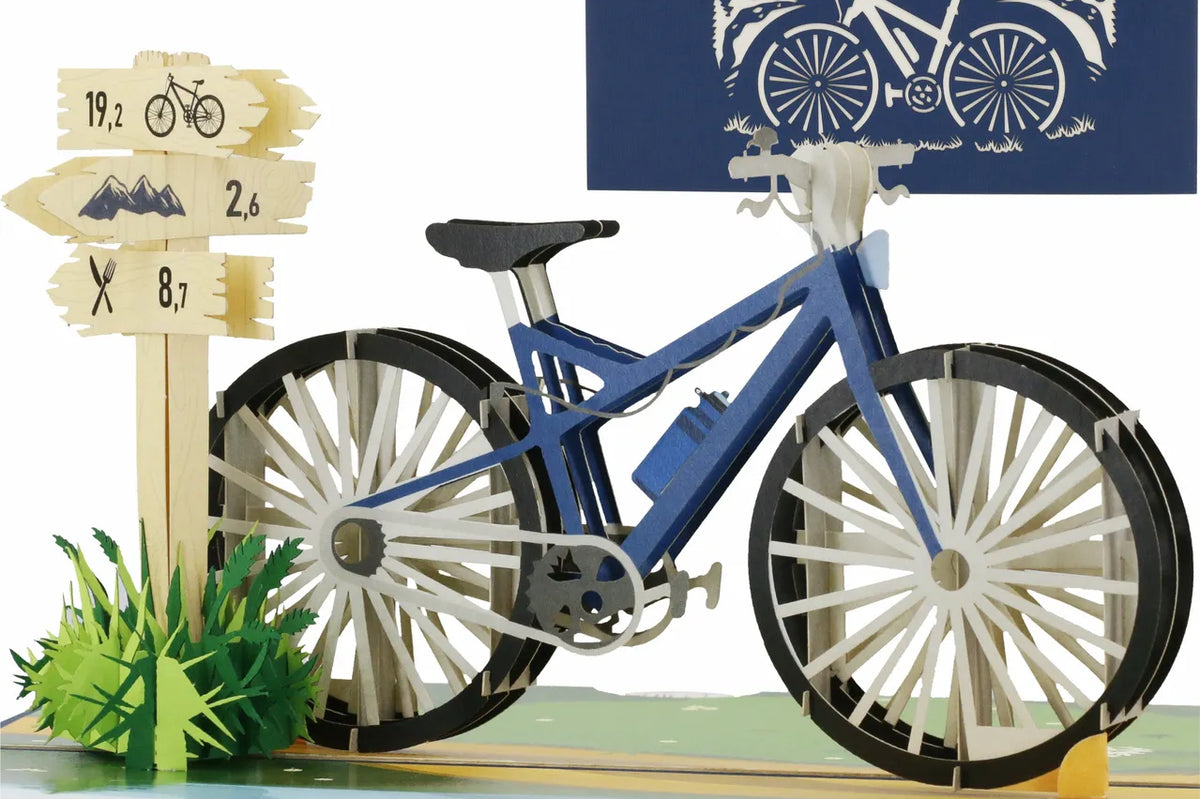 Pop Up Karte eines blauen Fahrrads mit zwei Wegweisern und einer Tafel mit Berglandschaft und Fahrradzeichnung im Hintergrund