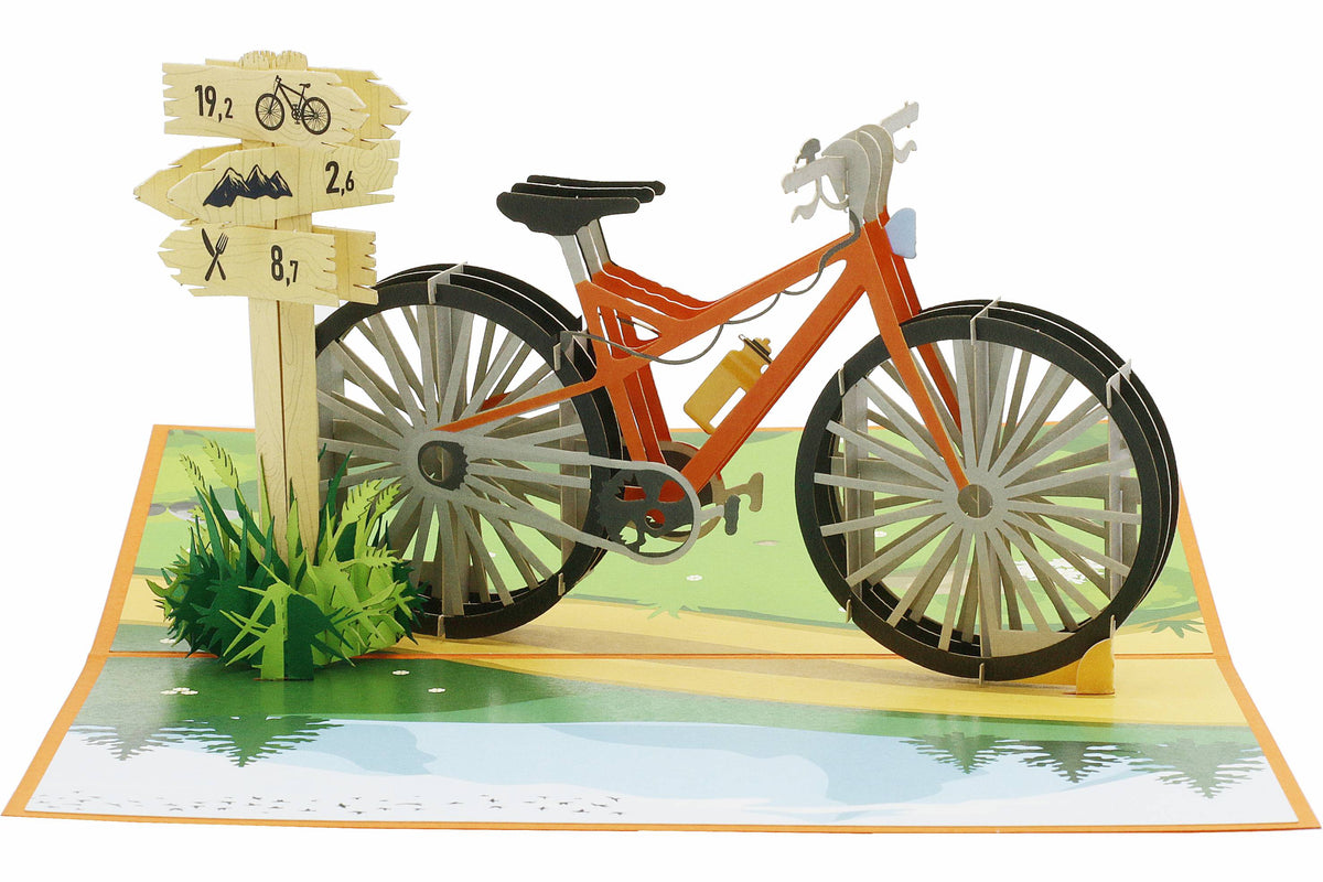 Aufgeklappte Pop-up-Karte mit orangem Fahrrad, Wegweiser und Wiese, frontal fotografiert.