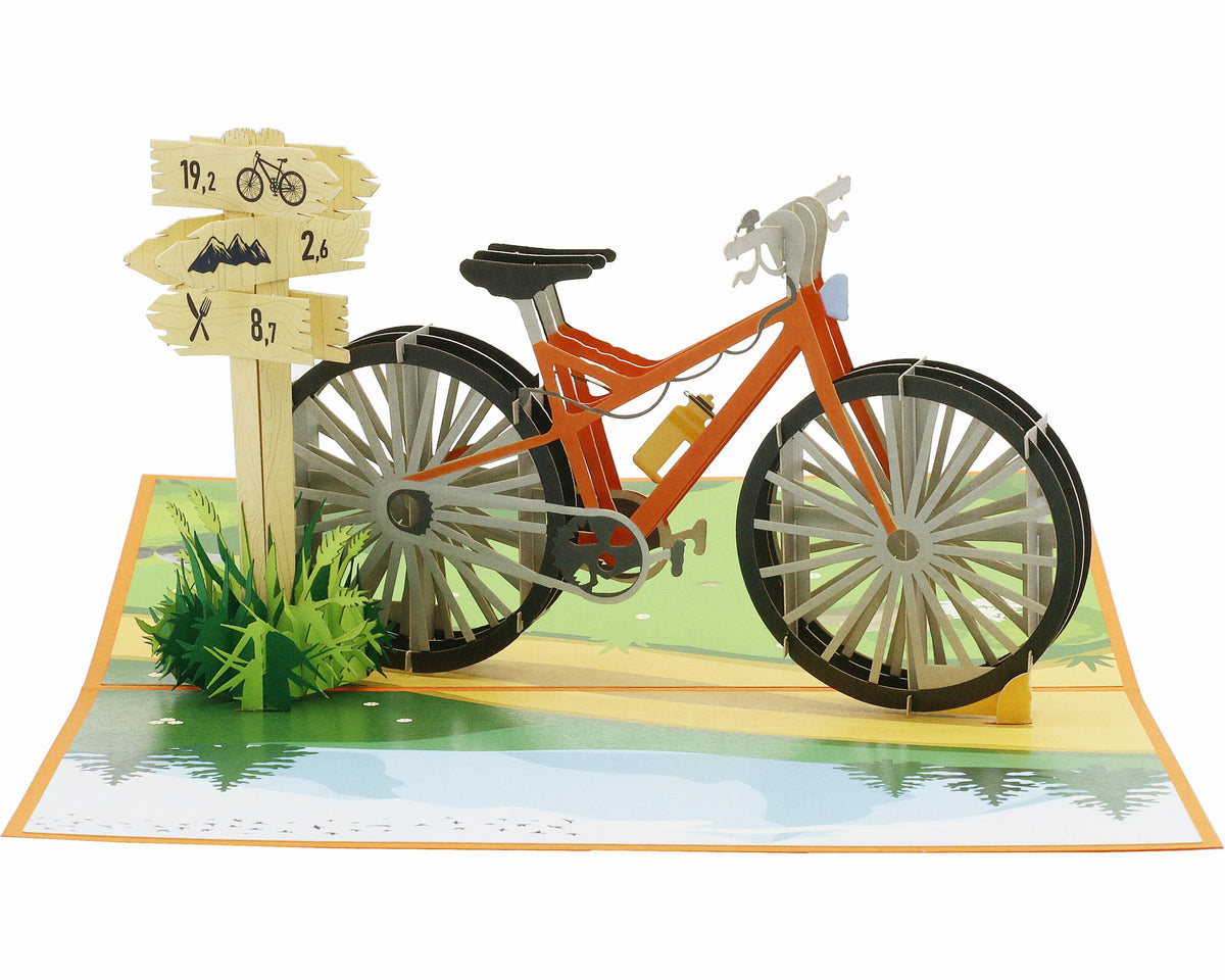 Aufgeklappte Pop-up-Karte mit orangem Fahrrad, Wegweiser und Wiese, frontal fotografiert.