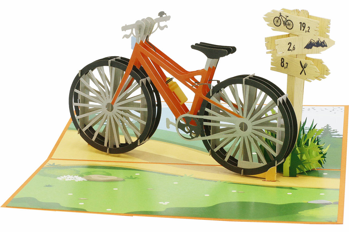 Aufgeklappte Pop-up-Karte mit orangem Fahrrad in einer Wiesenlandschaft, seitlich fotografiert.