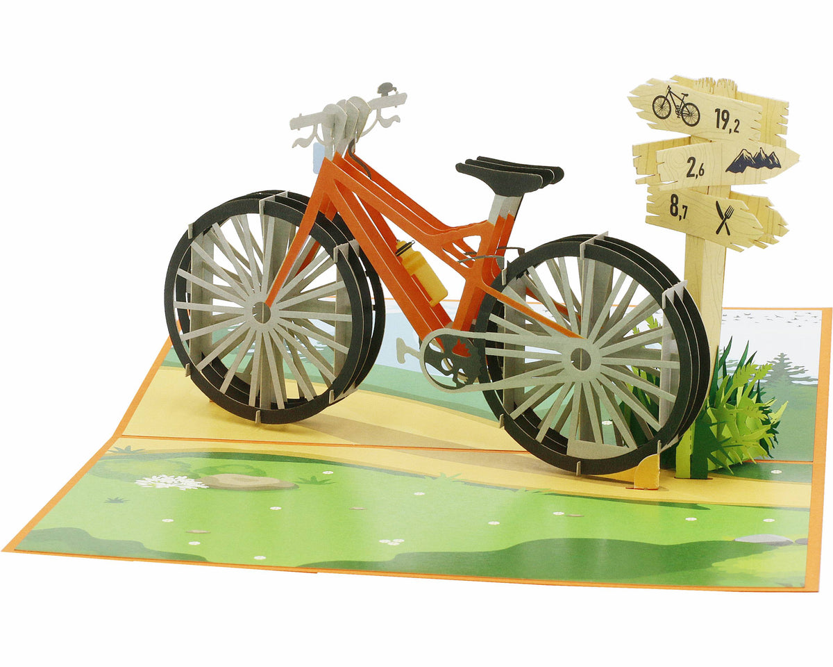 Aufgeklappte Pop-up-Karte mit orangem Fahrrad in einer Wiesenlandschaft, seitlich fotografiert.