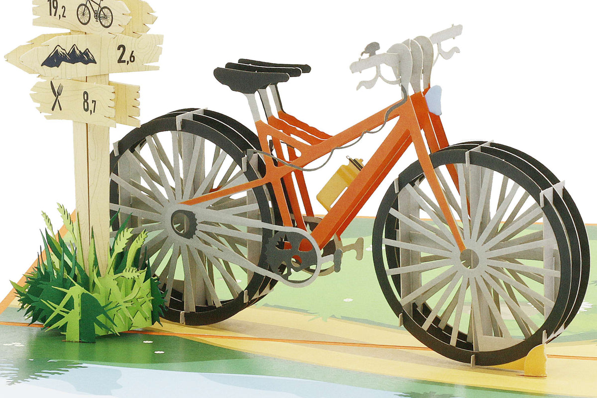 Aufgeklappte Pop-up-Karte mit einem orangen Fahrrad und einem Wegweiser mit Rad- und Wandersymbolen in einer Wiesenlandschaft.