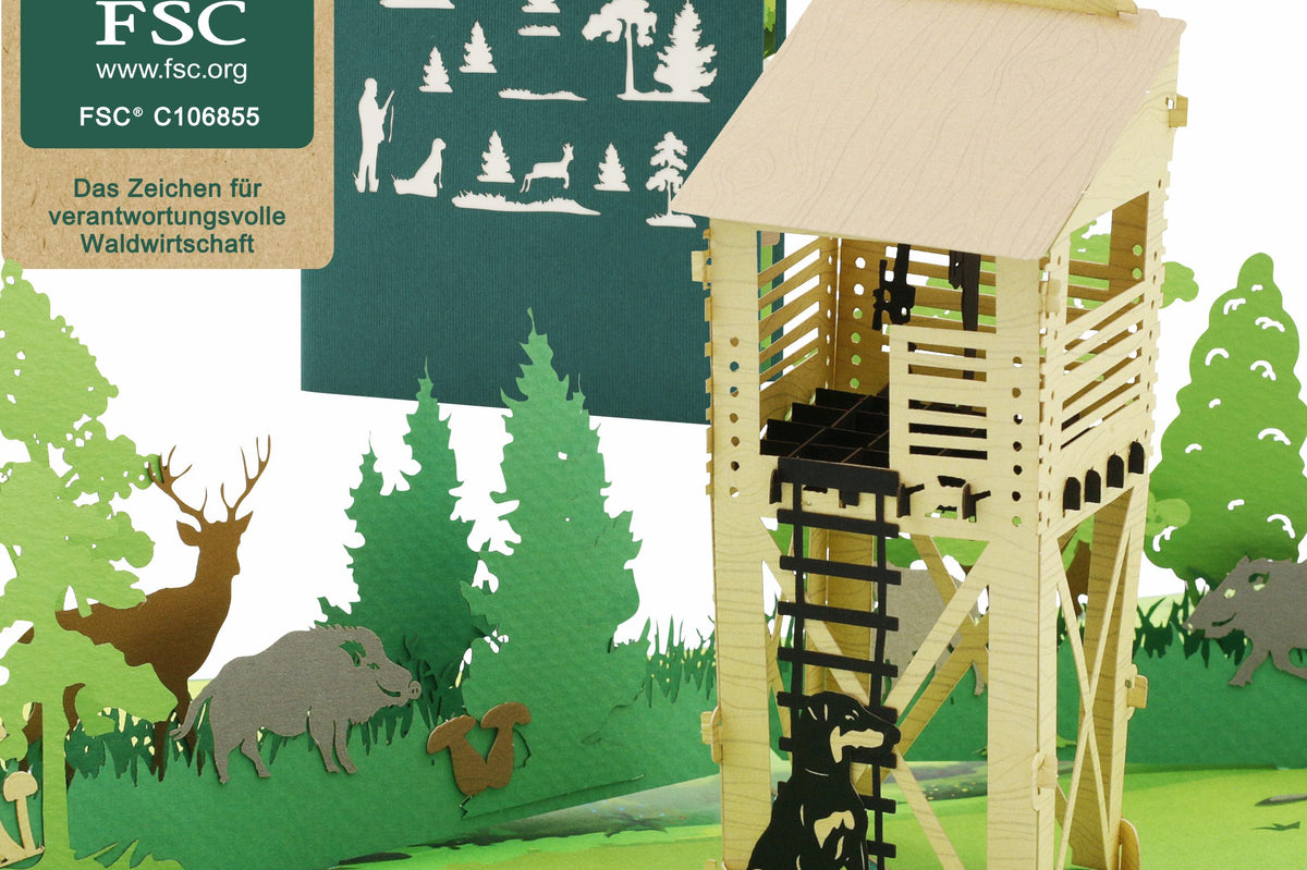 Aufgeklappte Pop-up-Karte mit einem hölzernen Hochsitz und Tieren in einer Waldlandschaft, im Hintergrund eine grüne Karte mit einem Lasercut von Wildtieren und Bäumen sowie ein FSC-Logo.