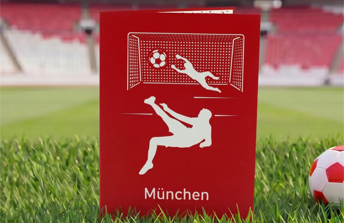 Rote Klappkarte mit Scherenschnitt eines Fußballspielers beim Fallrückzieher vor einem Tor und dem Schriftzug München auf Rasen im Stadion.