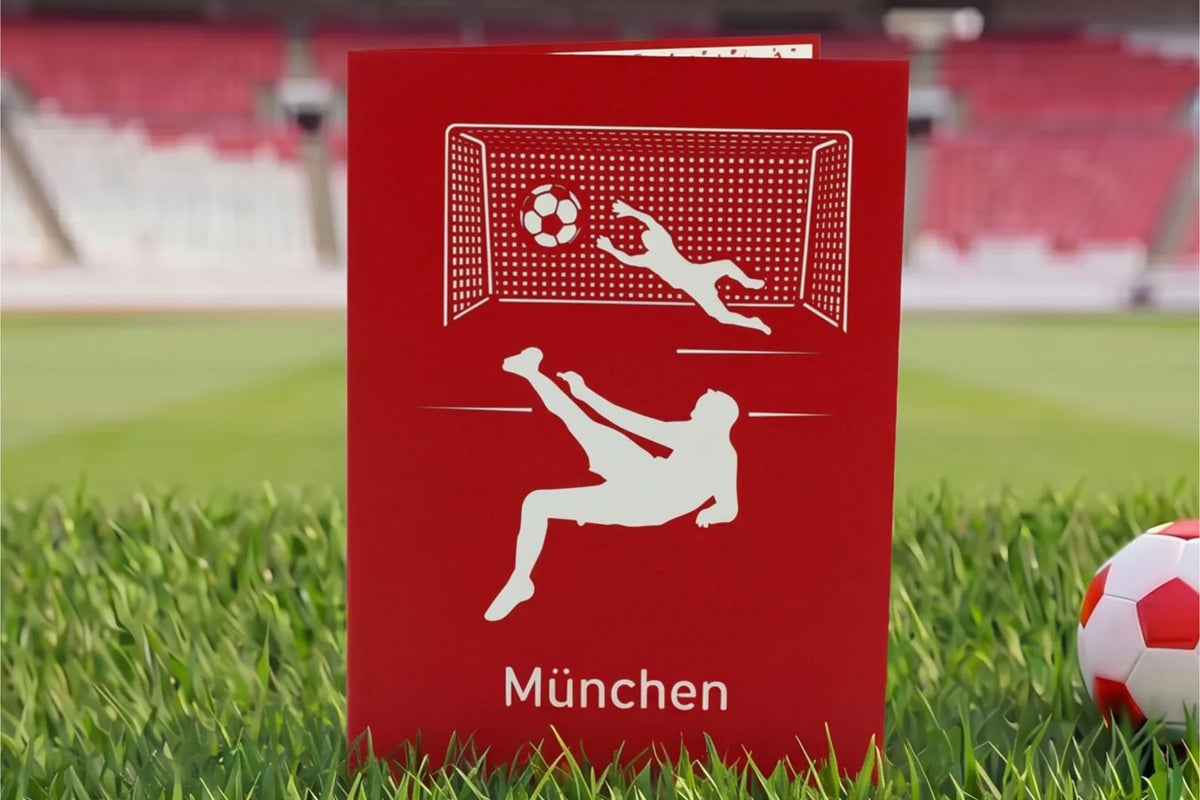 Rote Klappkarte mit Scherenschnitt eines Fußballspielers beim Fallrückzieher vor einem Tor und dem Schriftzug München auf Rasen im Stadion.