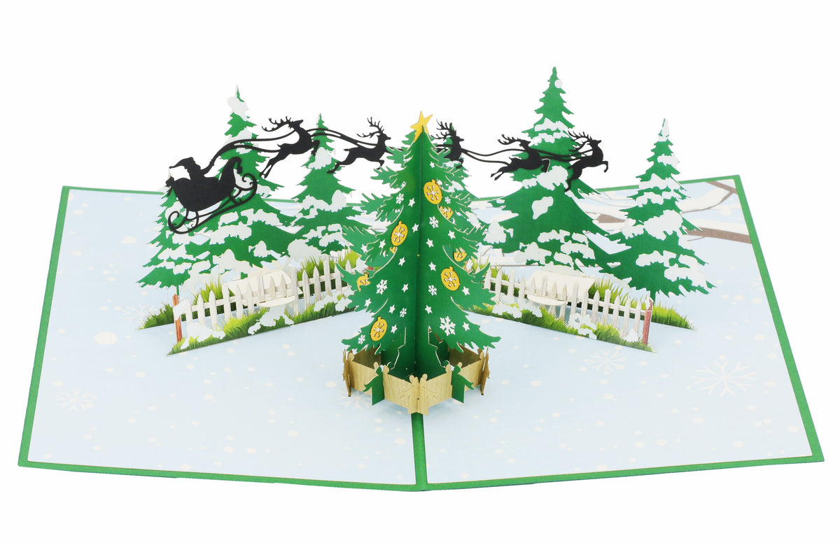 Aufgeklappte Pop-up-Karte mit einem geschmückten Tannenbaum, verschneiten Tannen und einem Rentierschlitten am Himmel.