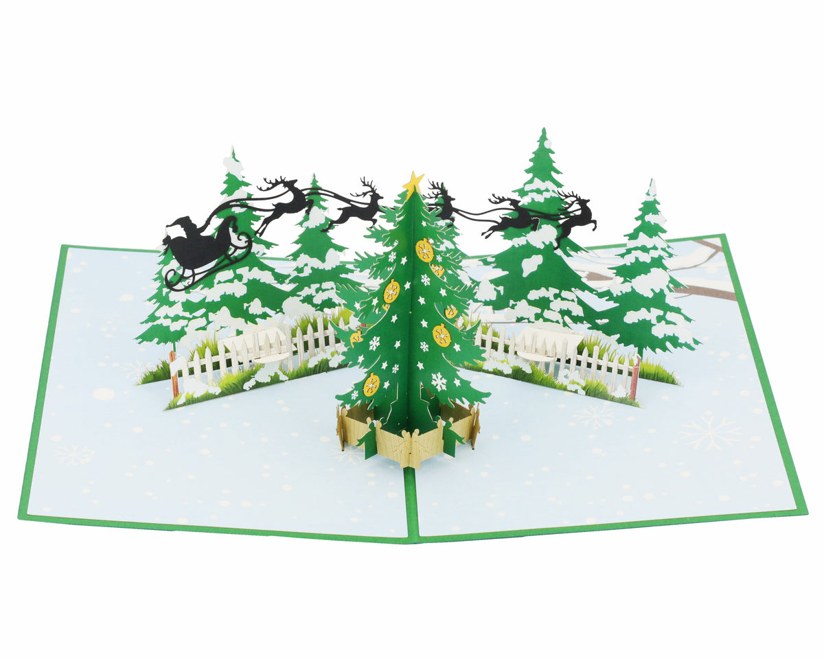 Aufgeklappte Pop-up-Karte mit einem geschmückten Tannenbaum, verschneiten Tannen und einem Rentierschlitten am Himmel.