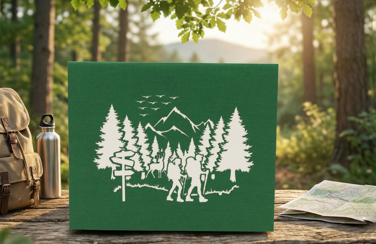 Grüne Klappkarte mit einem Wald, Bergen und 2 Wandersleuten in der Natur als Scherenschnitt. Die Pop Up Karte steht auf einer Bank in der Natur und im Hintergrund ist ein Wald.