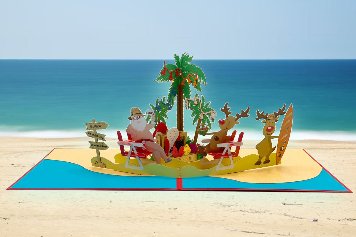 Pop-up-Karte mit Weihnachtsmann und Rentieren am Strand, aufgestellt vor einem echten Sandstrand mit Meer im Hintergrund.