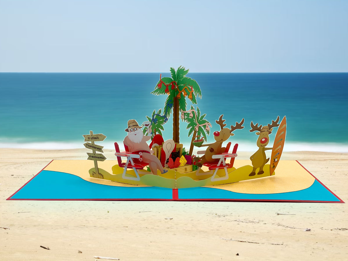 Pop-up-Karte mit Weihnachtsmann und Rentieren am Strand, aufgestellt vor einem echten Sandstrand mit Meer im Hintergrund.