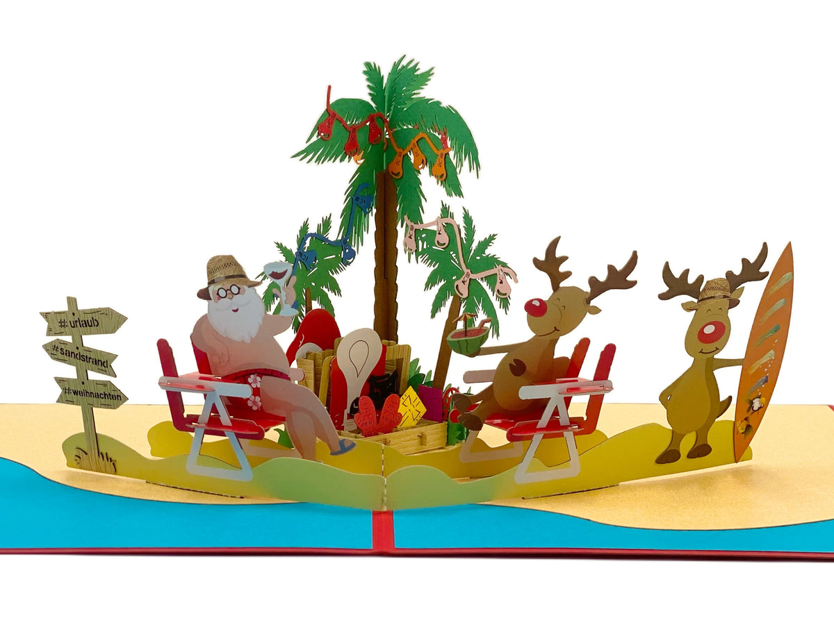 Nahaufnahme der aufgeklappten Pop-up-Karte: Weihnachtsmann im Liegestuhl mit Sonnenhut unter einer mit Christbaumkugeln geschmückten Palme, daneben zwei Rentiere mit Surfbrett am Sandstrand.