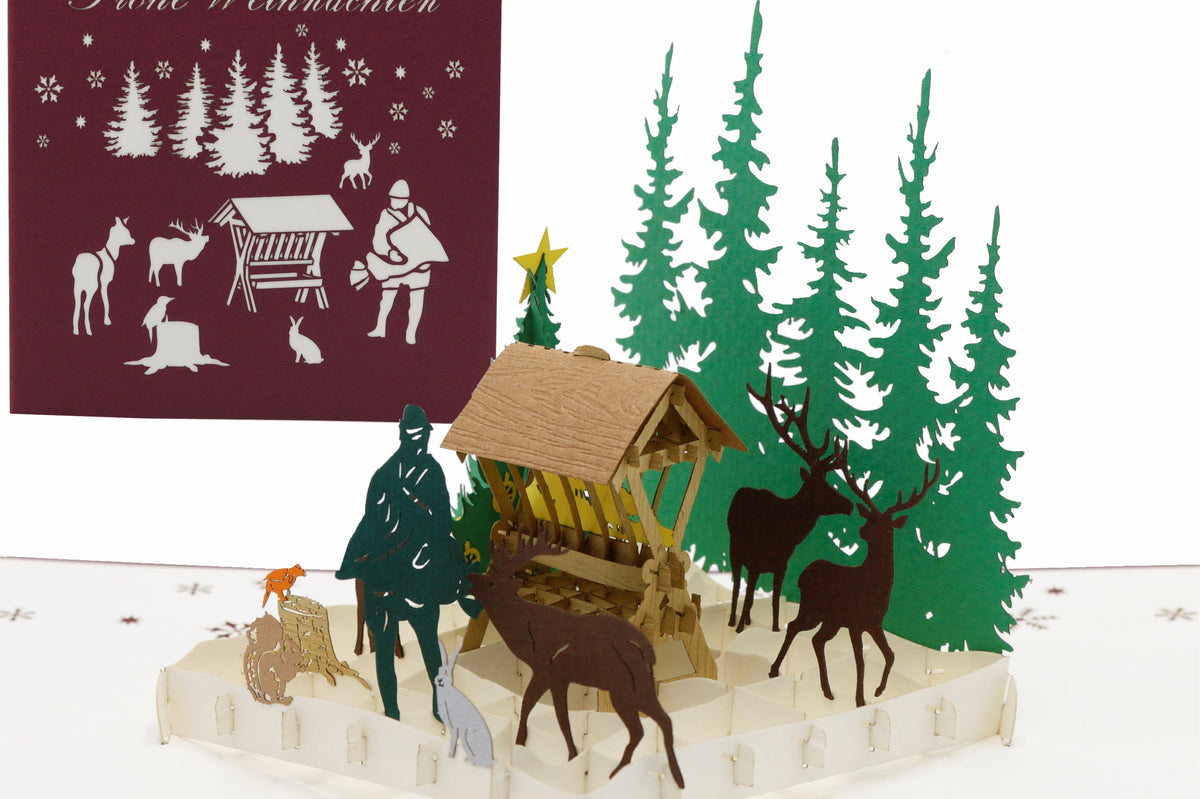 Aufgeklappte Pop-up-Karte mit Futterkrippe und Waldtieren, daneben die Kartenvorderseite mit dem Schriftzug Frohe Weihnachten.