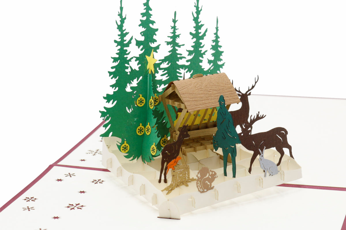 Aufgeklappte Pop-up-Karte mit Futterkrippe, Hirsch und Rehen im verschneiten Wald, seitlich fotografiert.