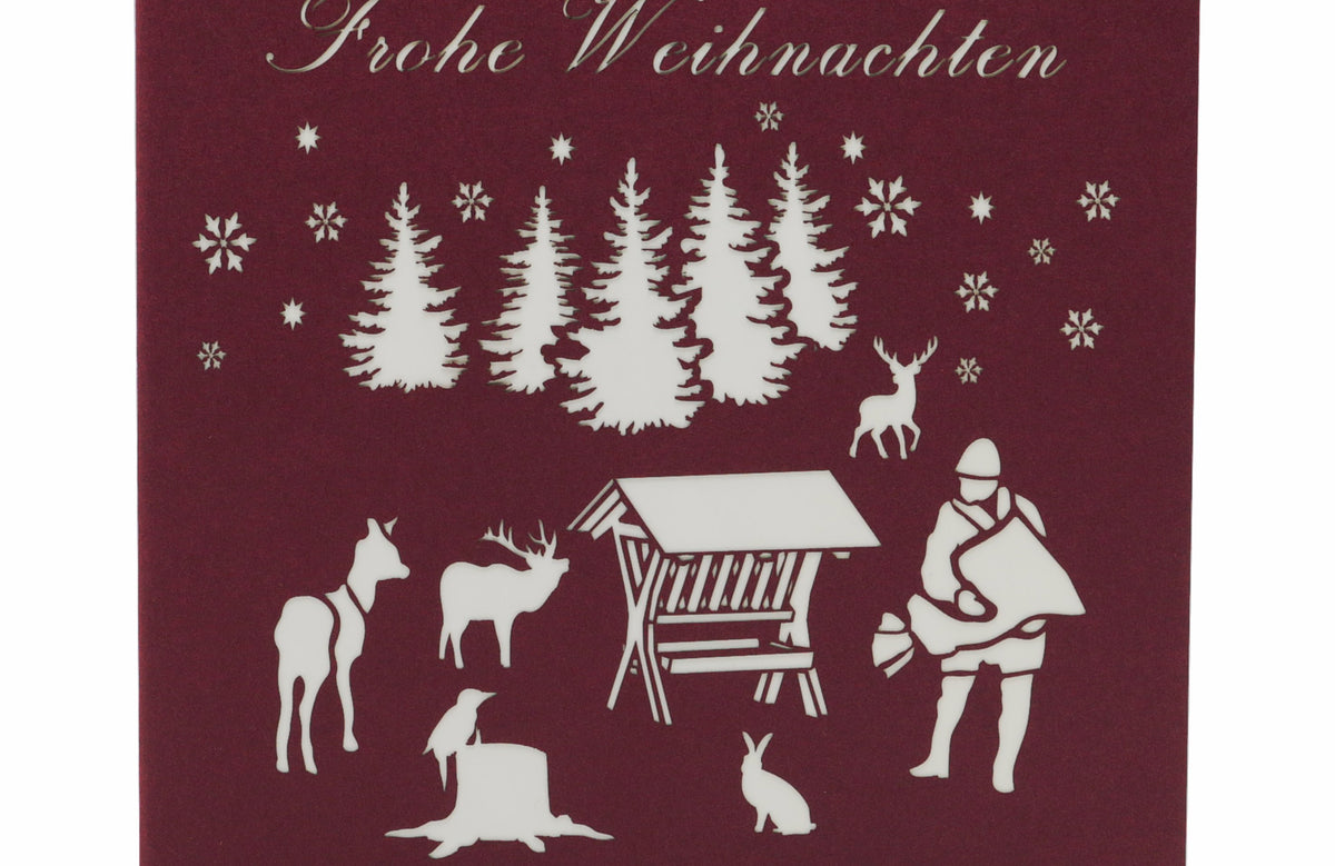 Dunkelrote Kartenvorderseite mit Futterkrippe, Jäger und Waldtieren als Scherenschnitt sowie dem Schriftzug Frohe Weihnachten.