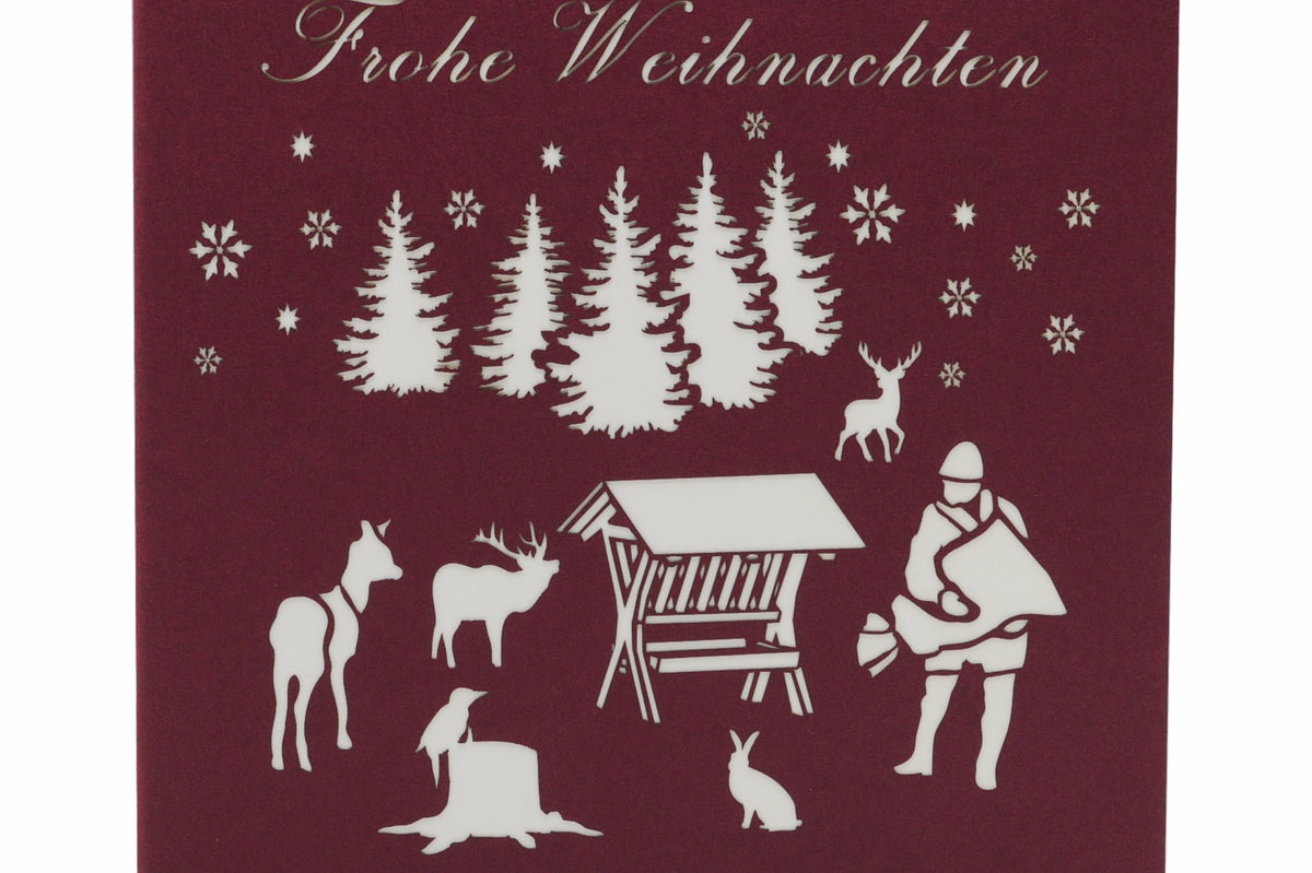 Dunkelrote Kartenvorderseite mit Futterkrippe, Jäger und Waldtieren als Scherenschnitt sowie dem Schriftzug Frohe Weihnachten.