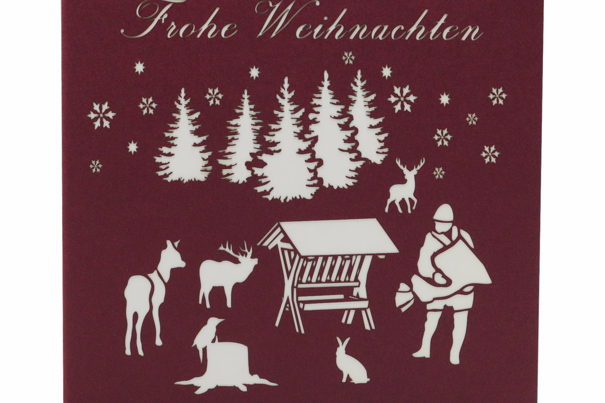 Dunkelrote Kartenvorderseite mit Futterkrippe, Jäger und Waldtieren als Scherenschnitt sowie dem Schriftzug Frohe Weihnachten.