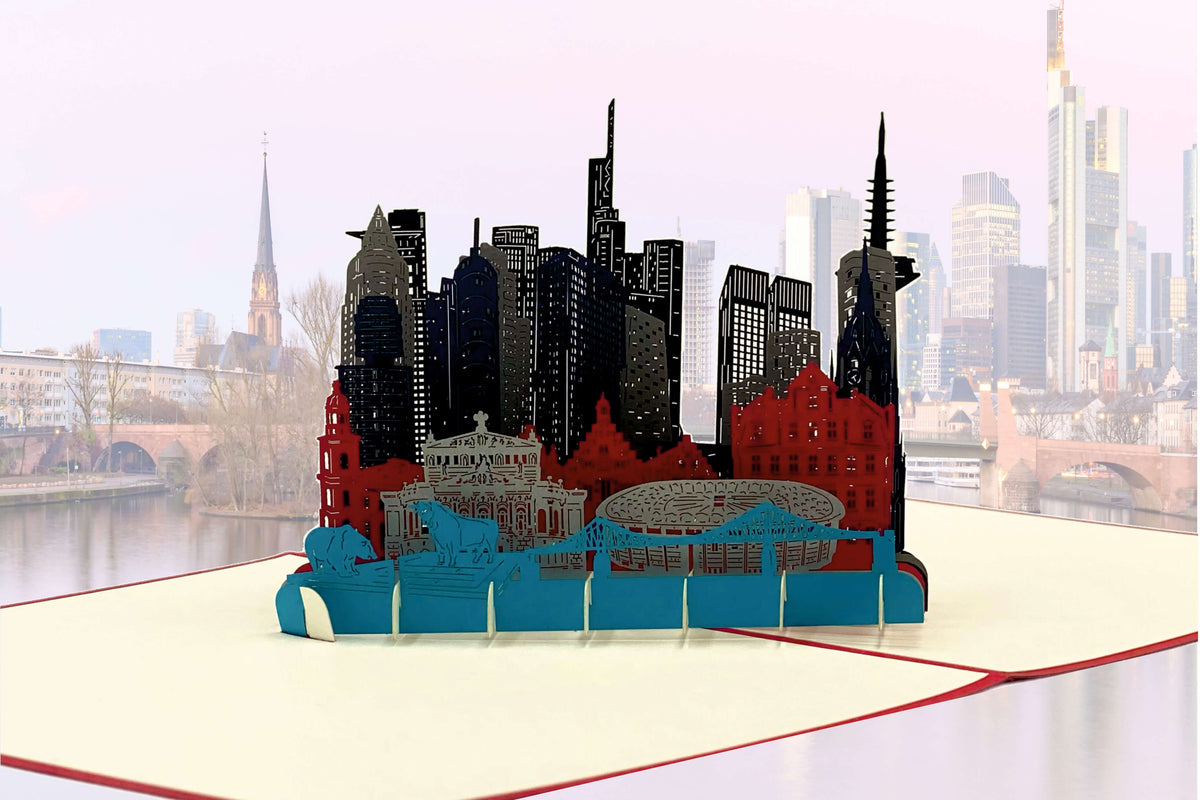 Aufgeklappte Pop-up-Karte mit der Skyline von Frankfurt vor einem echten Blick auf die Stadt am Main.