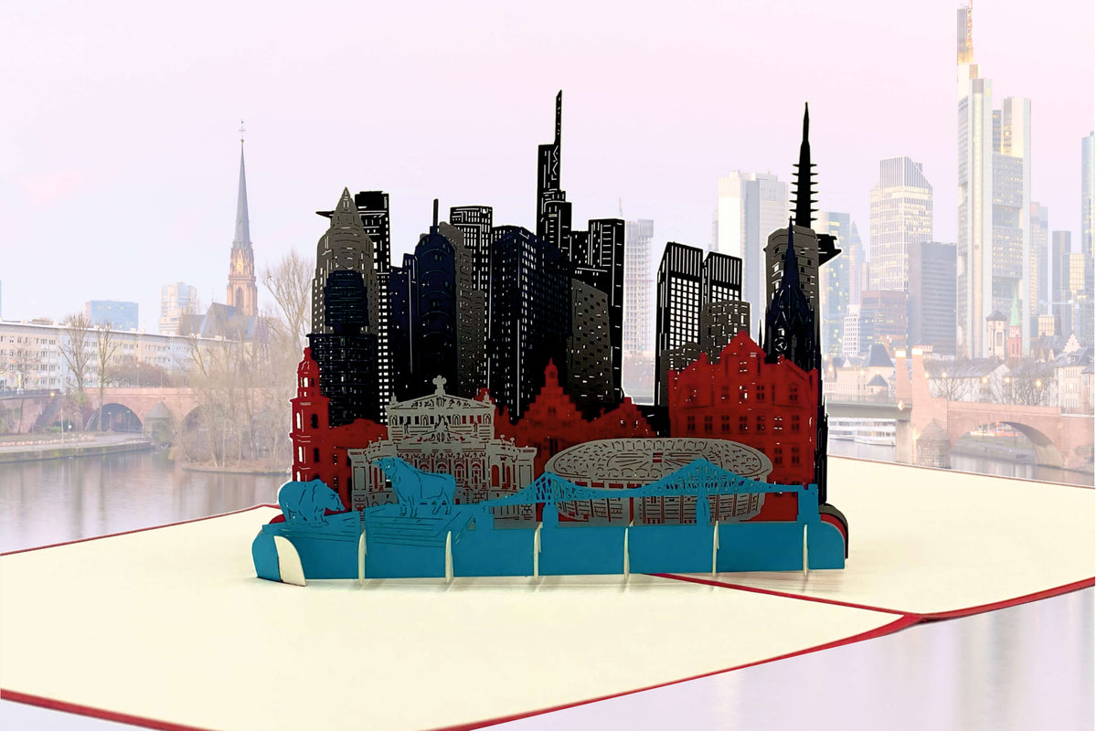 Aufgeklappte Pop-up-Karte mit der Skyline von Frankfurt vor einem echten Blick auf die Stadt am Main.