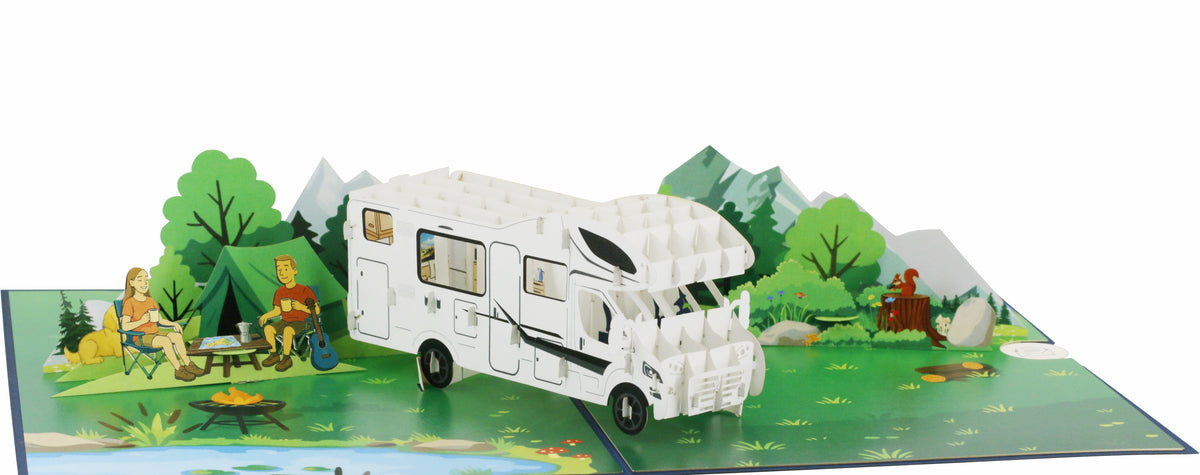 Aufgeklappte Pop-up-Karte mit weißem Wohnmobil, Bäumen und Bergen aus Papier, frontal fotografiert.