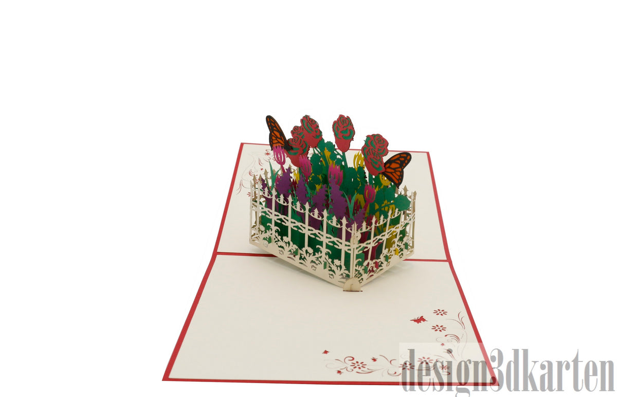 Aufgeklappte Pop-up-Karte mit rotem Rand: bunter Blumengarten hinter einem weißen Gartenzaun mit Schmetterlingen.
