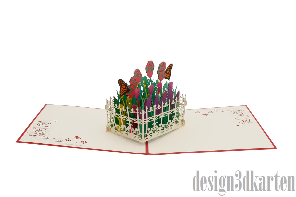 Aufgeklappte Pop-up-Karte mit Blumengarten, Gartenzaun und Schmetterlingen aus Papier, schräg fotografiert.