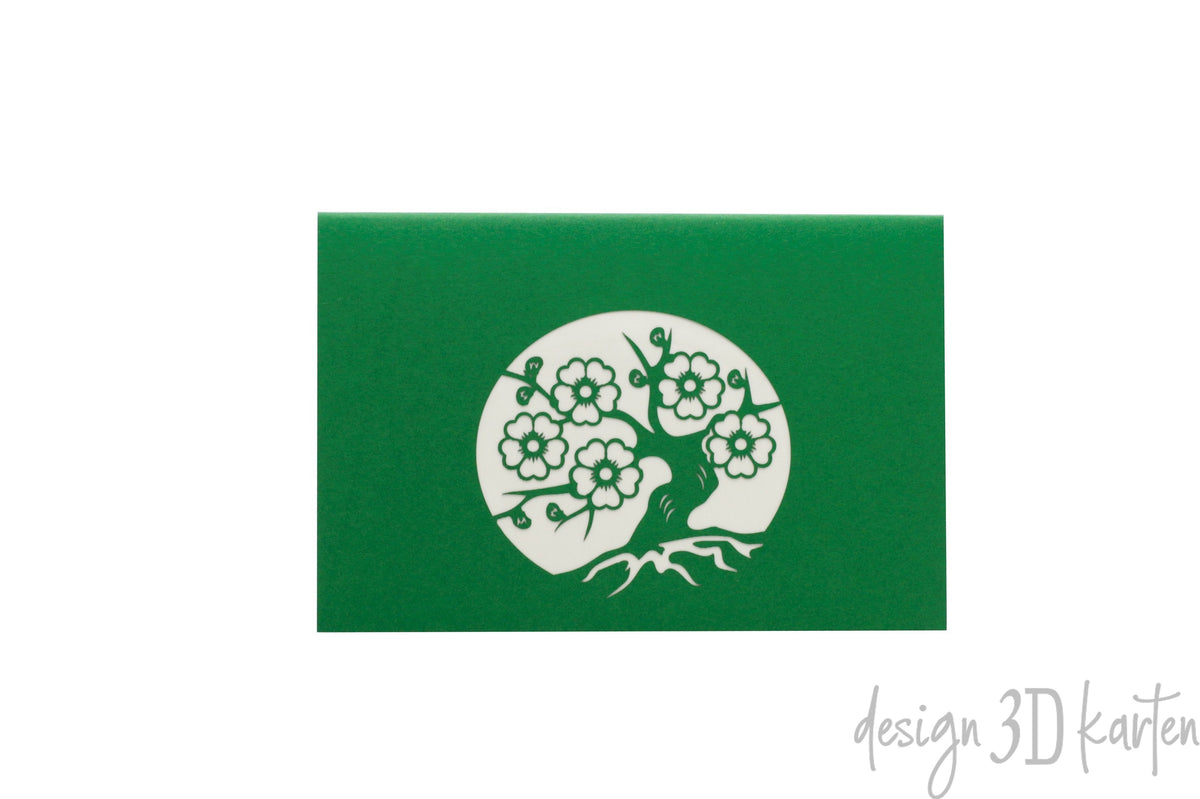 Grüne Kartenvorderseite mit einem Scherenschnitt: blühender Bonsai-Baum mit Wurzeln in einem Kreis.