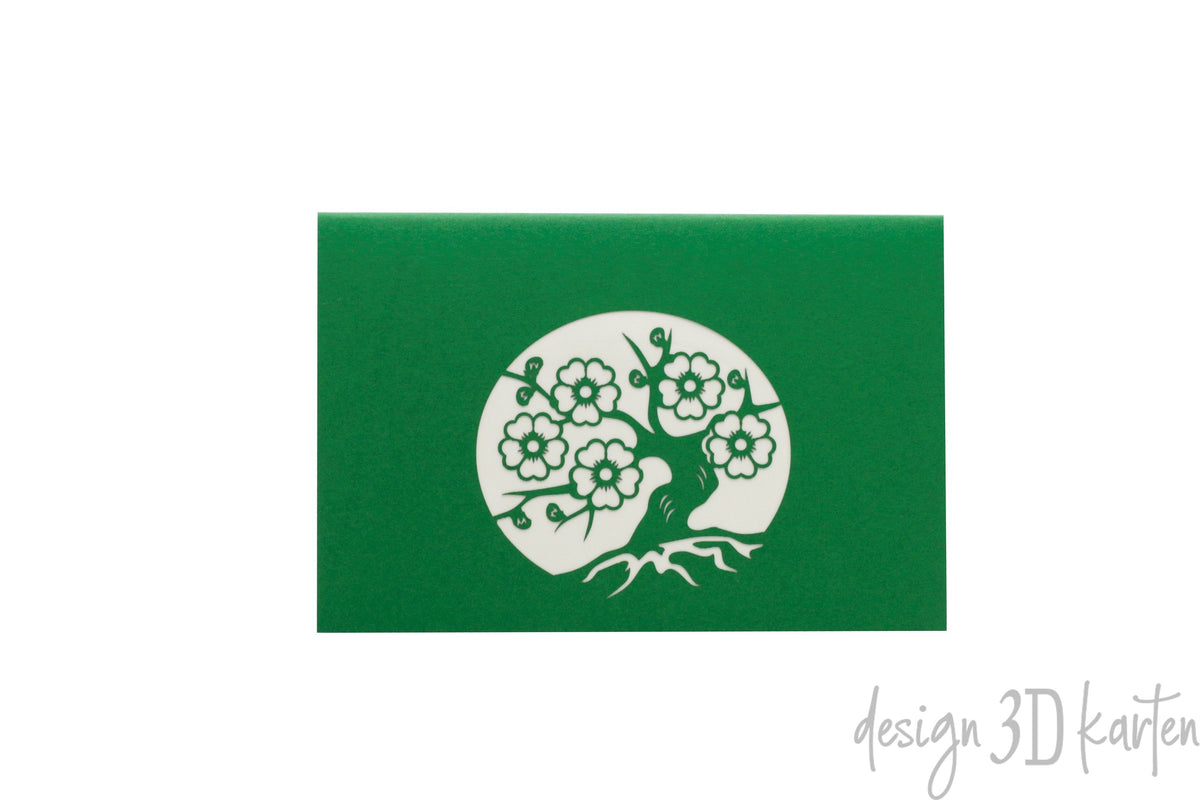 Grüne Kartenvorderseite mit einem Scherenschnitt: blühender Bonsai-Baum mit Wurzeln in einem Kreis.