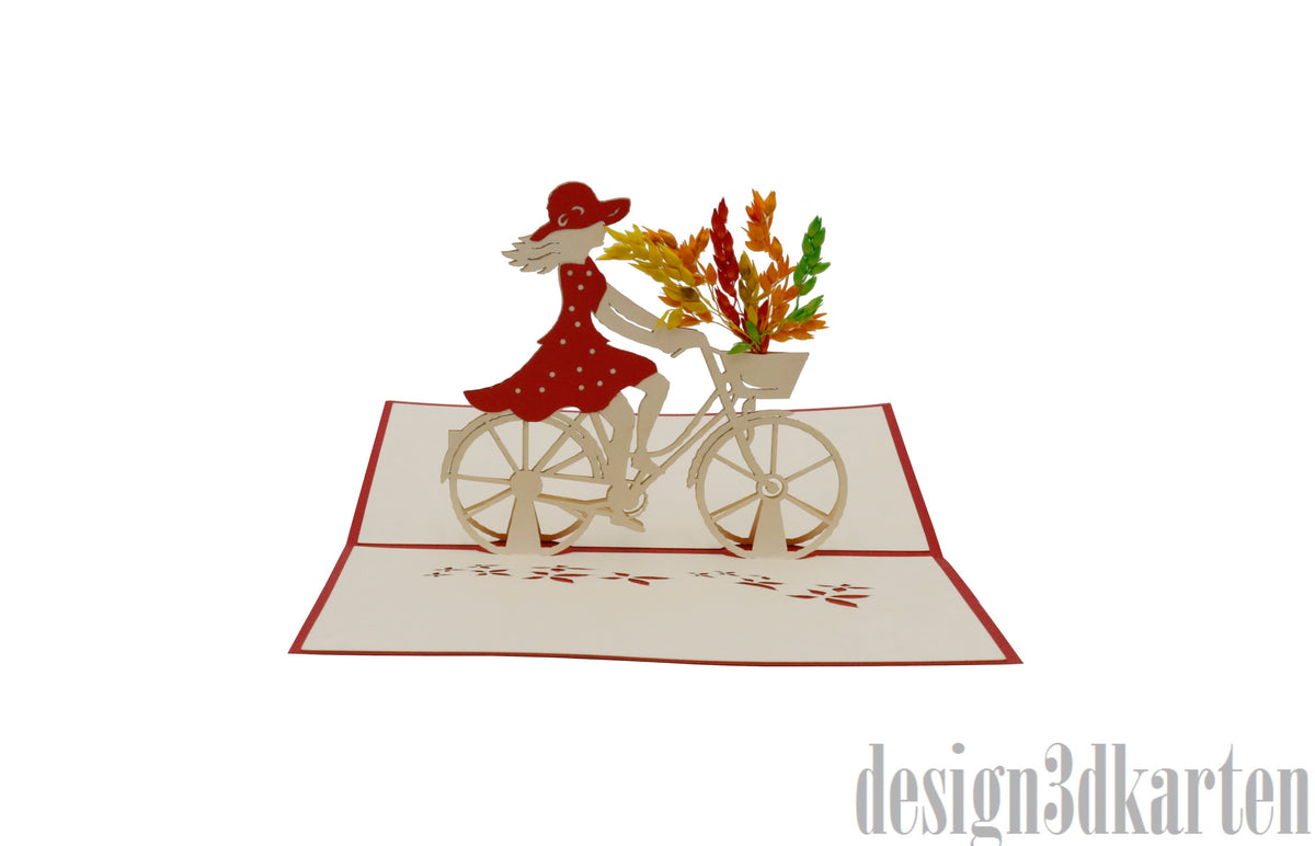 Aufgeklappte Pop-up-Karte mit rotem Rand: eine Frau im roten Kleid mit Hut fährt ein Damenfahrrad mit Blumenkorb.