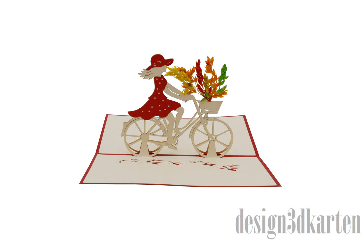 Aufgeklappte Pop-up-Karte mit rotem Rand: eine Frau im roten Kleid mit Hut fährt ein Damenfahrrad mit Blumenkorb.