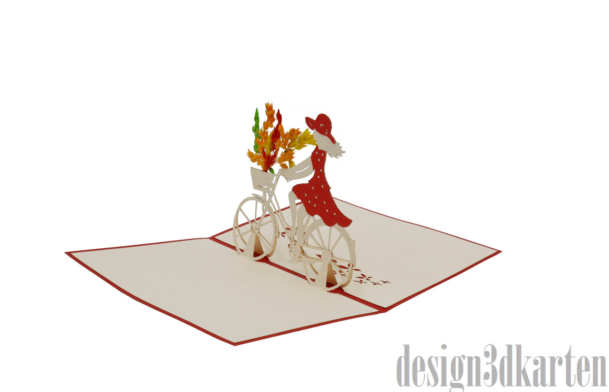 Aufgeklappte Pop-up-Karte mit Radfahrerin und Blumenkorb, schräg von der Seite fotografiert.