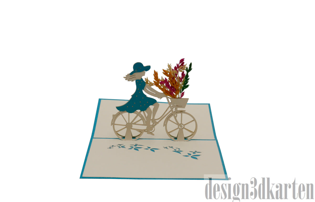 Aufgeklappte Pop-up-Karte mit türkisem Rand: eine Radfahrerin im blauen Kleid mit Blumenkorb am Fahrrad.