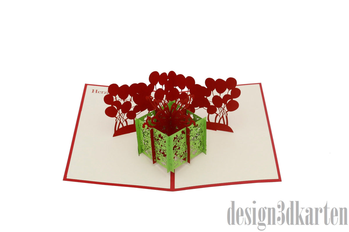 Aufgeklappte Pop-up-Karte mit grüner Geschenkbox und roten Luftballons, frontal fotografiert.