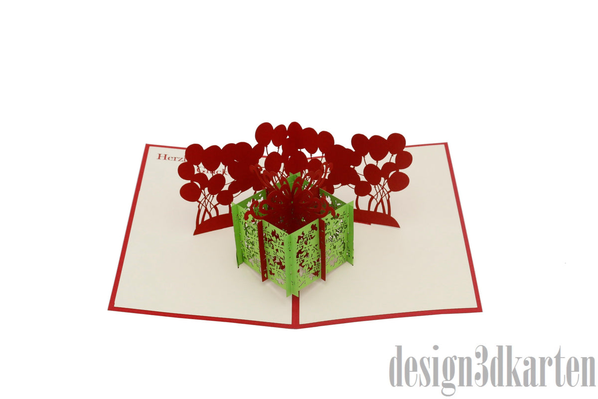 Aufgeklappte Pop-up-Karte mit grüner Geschenkbox und roten Luftballons, frontal fotografiert.