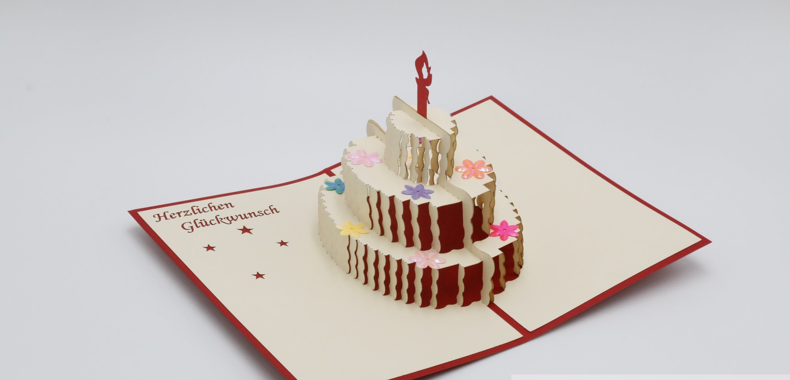 Aufgeklappte cremefarbene Pop-up-Karte mit roter Kante und einer dreistöckigen Geburtstagstorte mit Kerze, daneben der Schriftzug Herzlichen Glückwunsch.