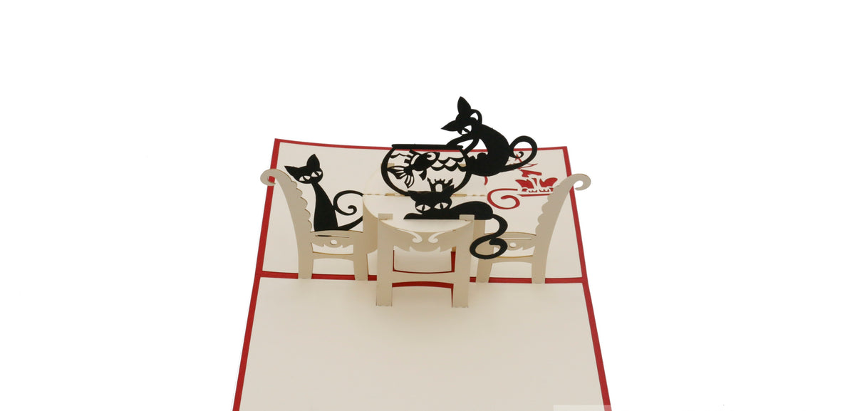 Aufgeklappte Pop-up-Karte mit rotem Rand: zwei schwarze Katzen an einem gedeckten Tisch mit Goldfischglas.