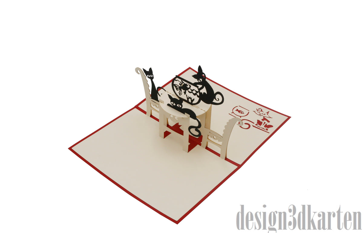 Aufgeklappte Pop-up-Karte mit zwei schwarzen Katzen am Tisch, schräg von der Seite fotografiert.