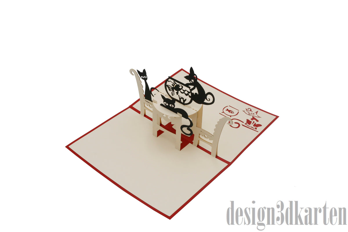 Aufgeklappte Pop-up-Karte mit zwei schwarzen Katzen am Tisch, schräg von der Seite fotografiert.