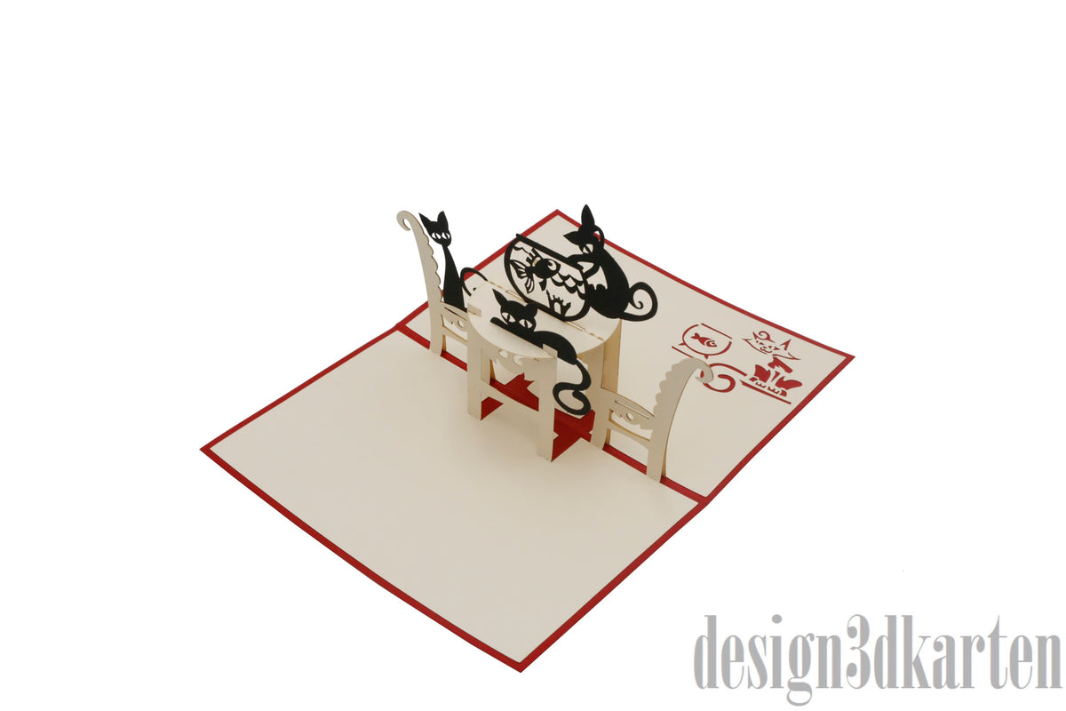 Aufgeklappte Pop-up-Karte mit zwei schwarzen Katzen am Tisch, schräg von der Seite fotografiert.