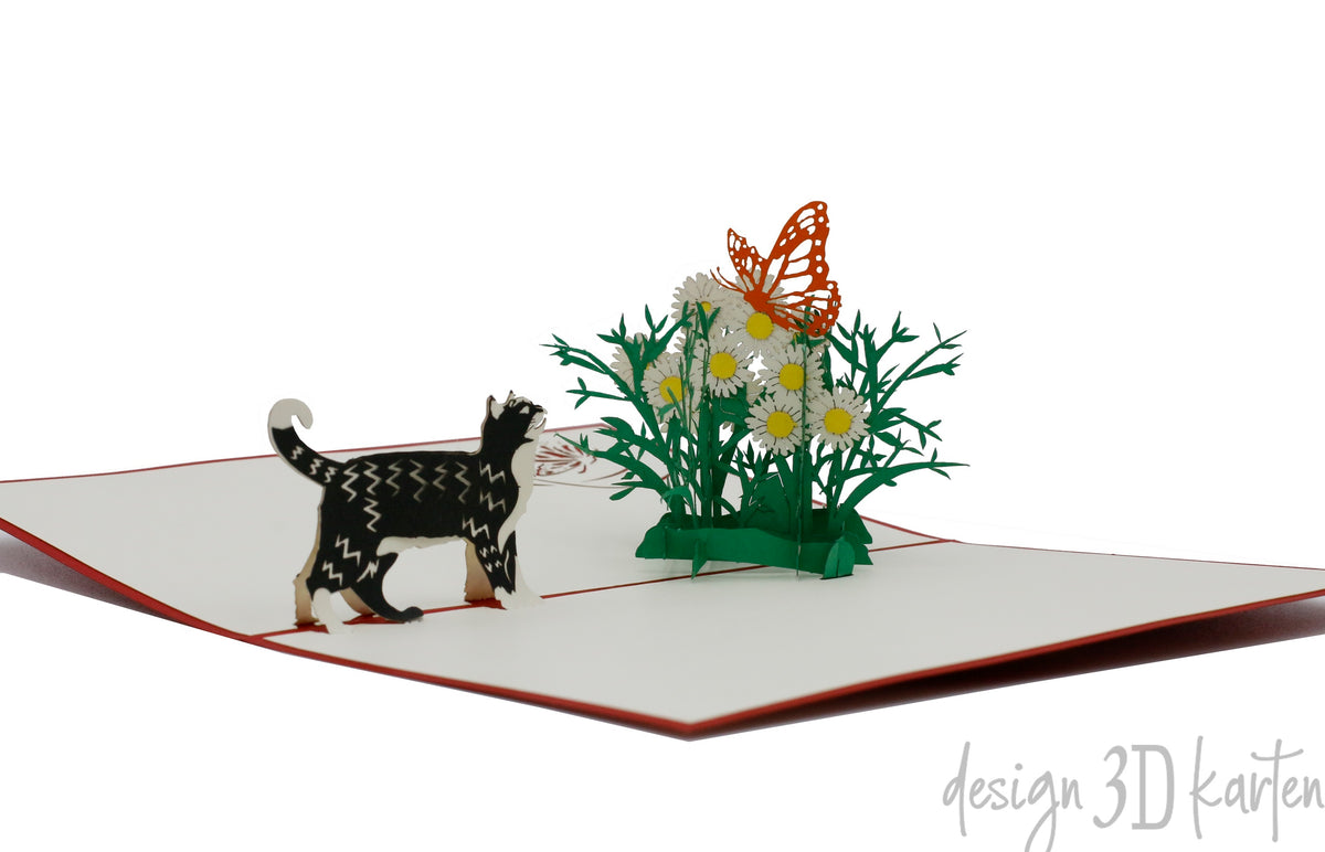 Aufgeklappte Pop-up-Karte mit rotem Rand: eine getigerte Katze läuft auf eine Wiese mit Margeriten und einem orangen Schmetterling zu.