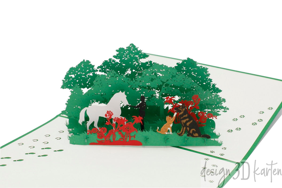Aufgeklappte Pop-up-Karte mit grünem Rand: ein weißes Pferd, ein Hund und Menschen in einer grünen Waldlandschaft mit roten Blüten.