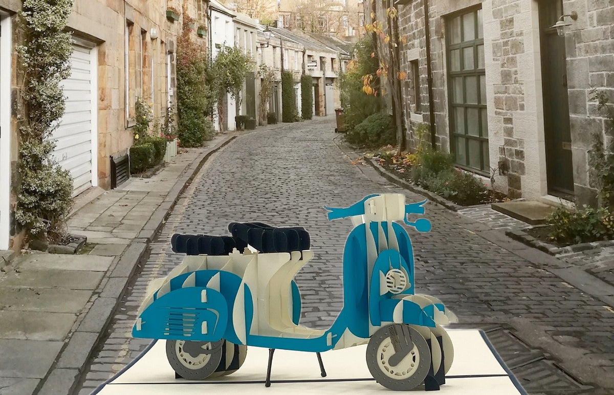 Pop-up-Karte mit blauer Vespa, aufgestellt in einer gepflasterten Altstadtgasse.