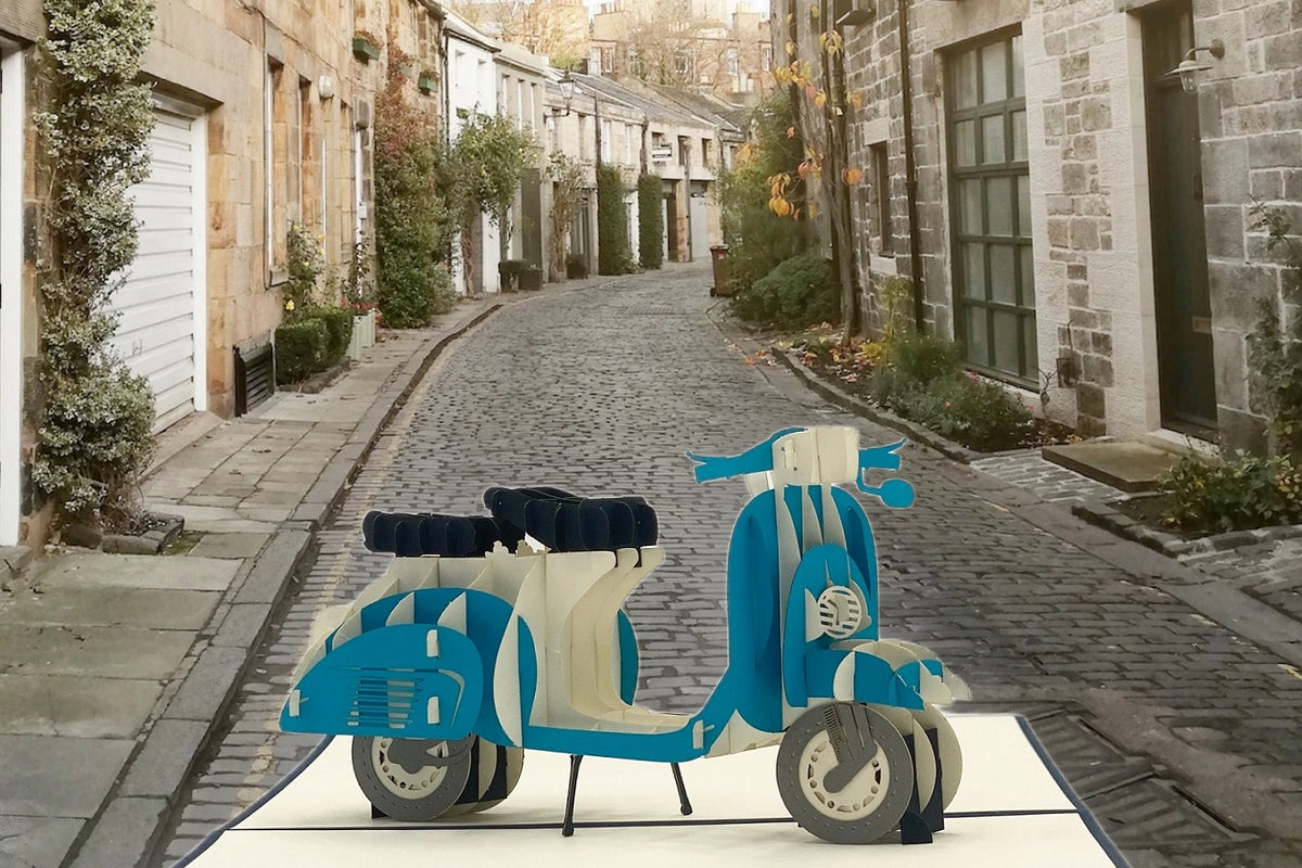 Pop-up-Karte mit blauer Vespa, aufgestellt in einer gepflasterten Altstadtgasse.