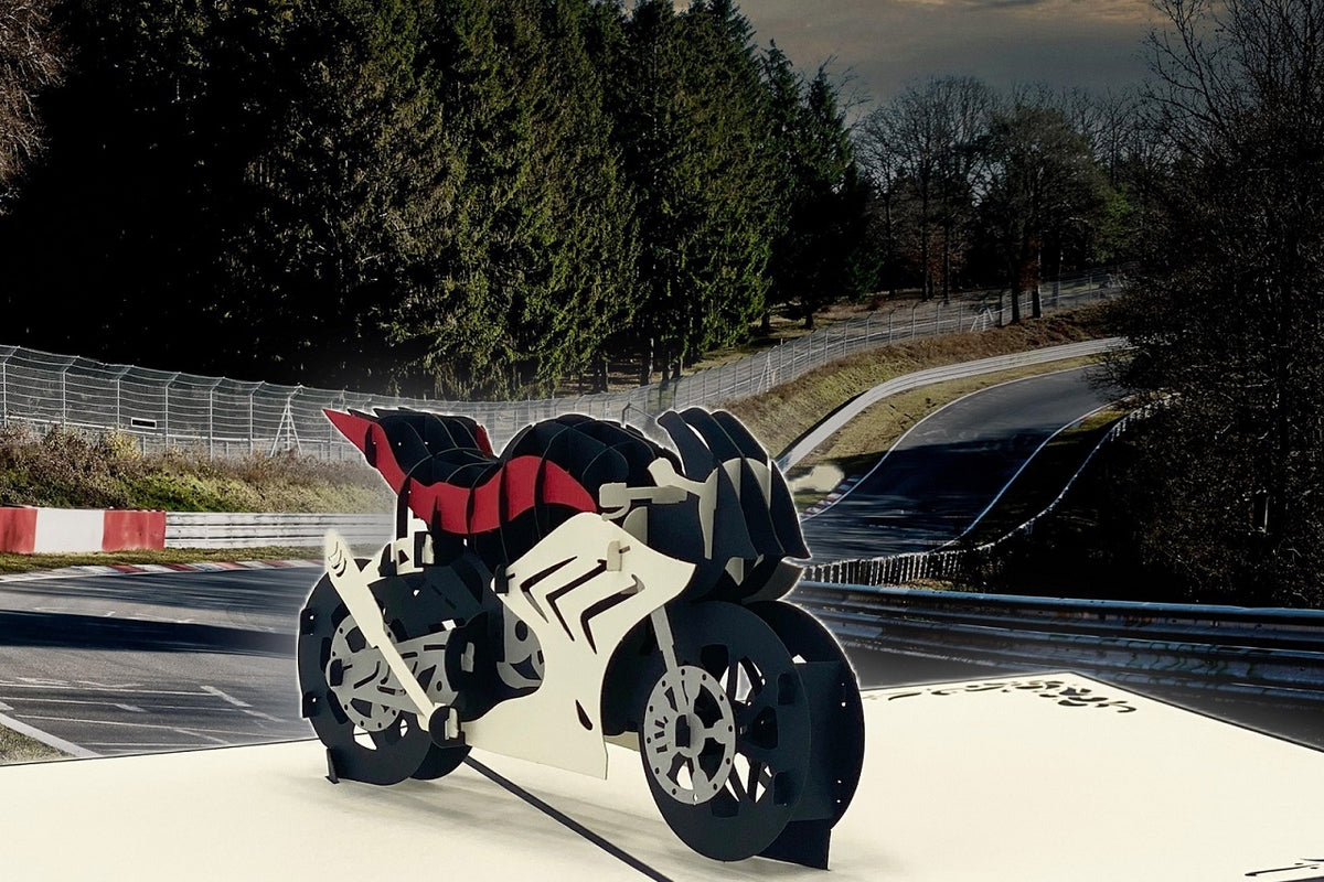 Pop-up-Karte mit Rennmotorrad, aufgestellt vor einer echten Rennstrecke mit Leitplanken und Wald.